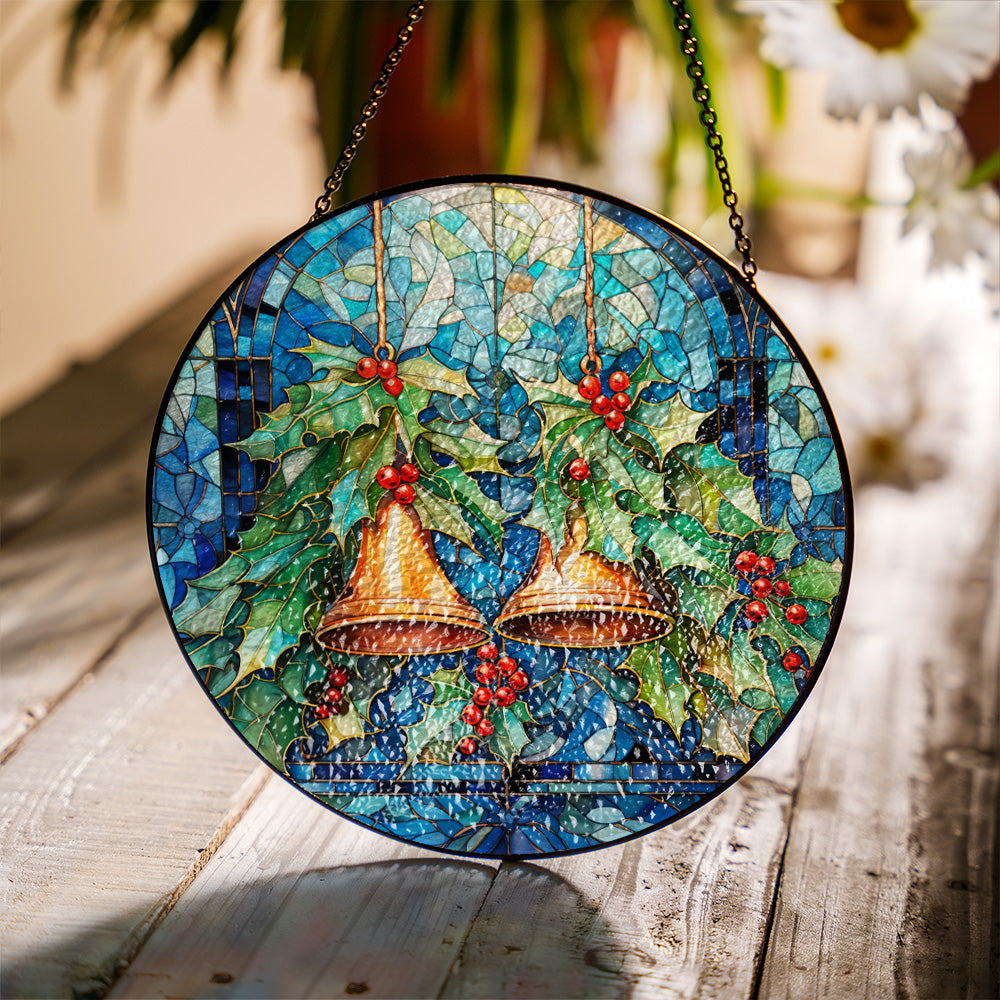 Round stained glass hanging ornament featuring two gold bells, green holly leaves, and red berries on a blue mosaic background, suspended by chains over a rustic wooden surface. Christmas bells stained glass suncatcher hanging in a sunlit window