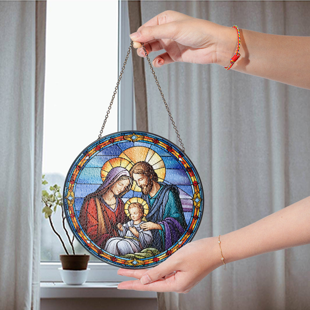 A round stained-glass-style hanging ornament depicting the Holy Family, held by two hands in front of a window with curtains. 